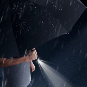 Person holding an open black umbrella with a flashlight in the handle, illumination falling rain droplets in the dark. Demonstrating umbrella flashlight feature.
