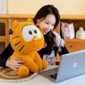 Smiling woman with long hair holding a large orange Garfield plush toy while looking at a silver laptop on a table.