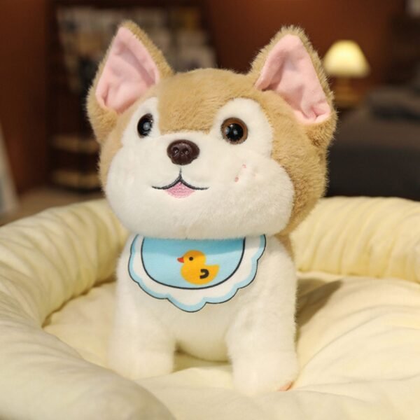 An adorable light brown and white husky puppy plush toy with pink ears, black eyes and a blue bib with a yellow duck on it, sitting in a soft light colored bed.