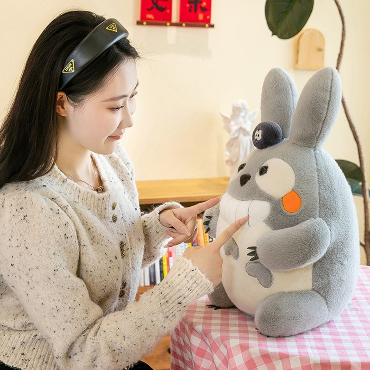 Young woman interacting with a large, grey, fuzzy plush toy resembling a rabbit with a slight grin and a black soot sprite on its head, for wholesale inventory.