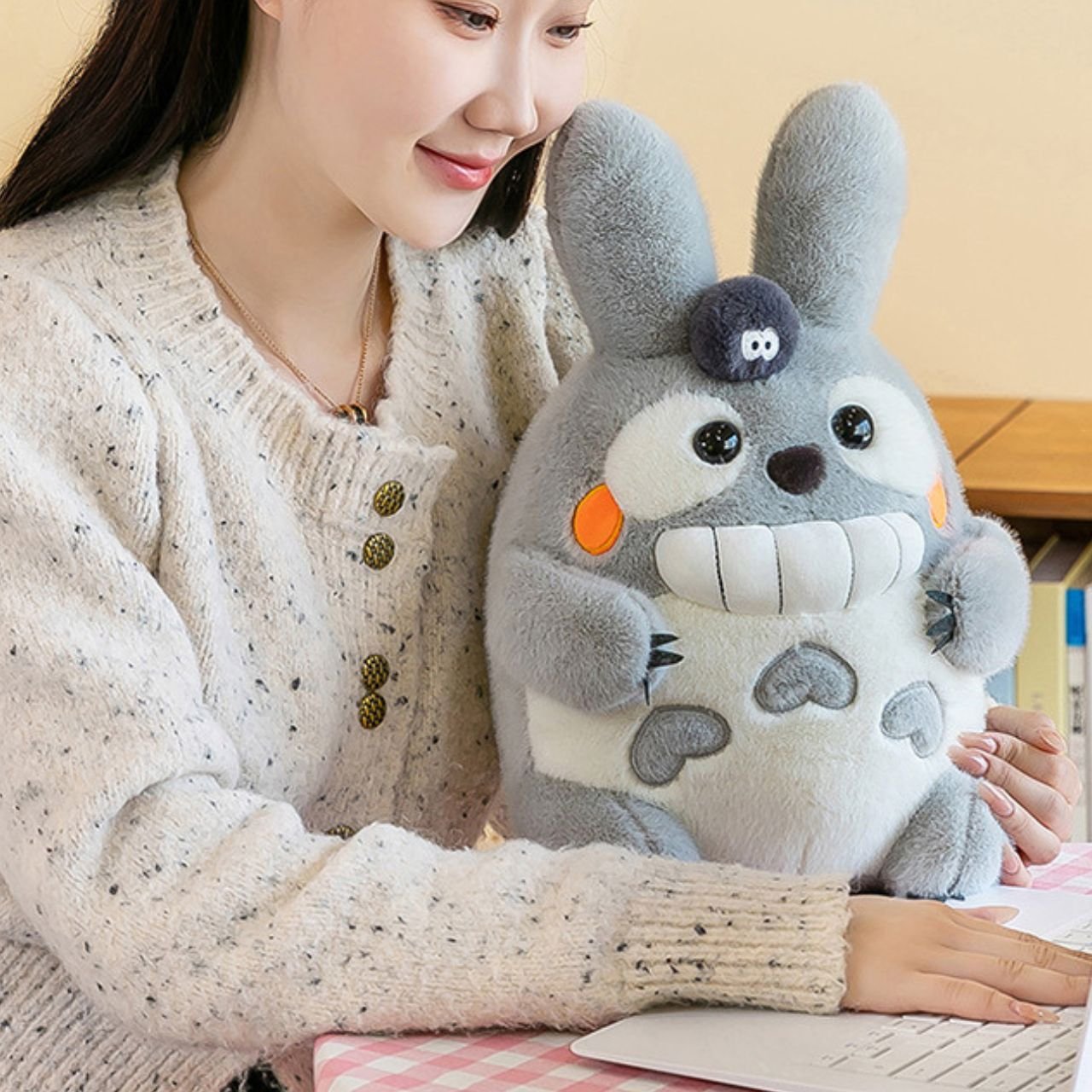 Smiling young woman wearing a cream cardigan, hugging a large, soft grey plush toy resembling Totoro with white chest and large ears, while sitting at a desk with a laptop.
