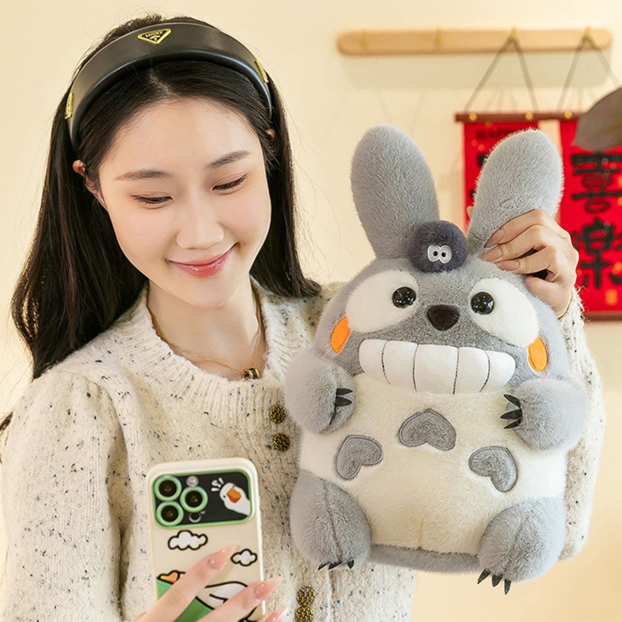 Smiling young woman holding a large, grey fantasy rabbit plush toy with a soot sprite, while looking at her smartphone with a whimsical case; ideal for wholesale product presentation.