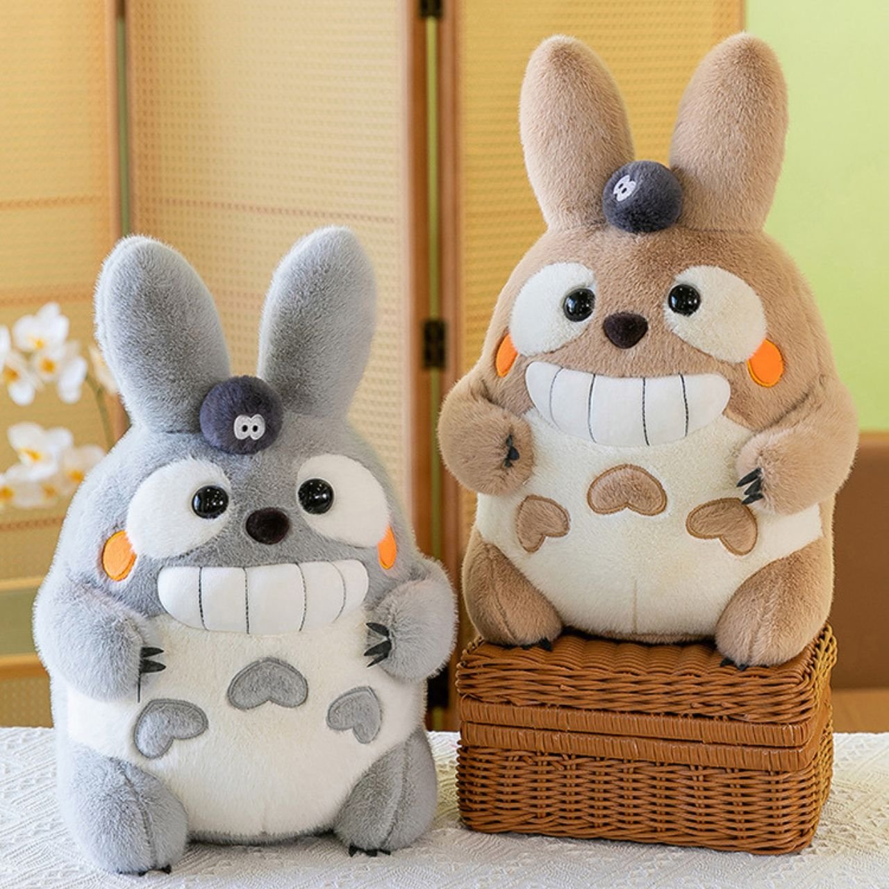 Two large, smiling plush rabbit toys, one grey and one brown resting on a wicker basket, both featuring a small black soot sprite; ideal for wholesale soft toy inventory.