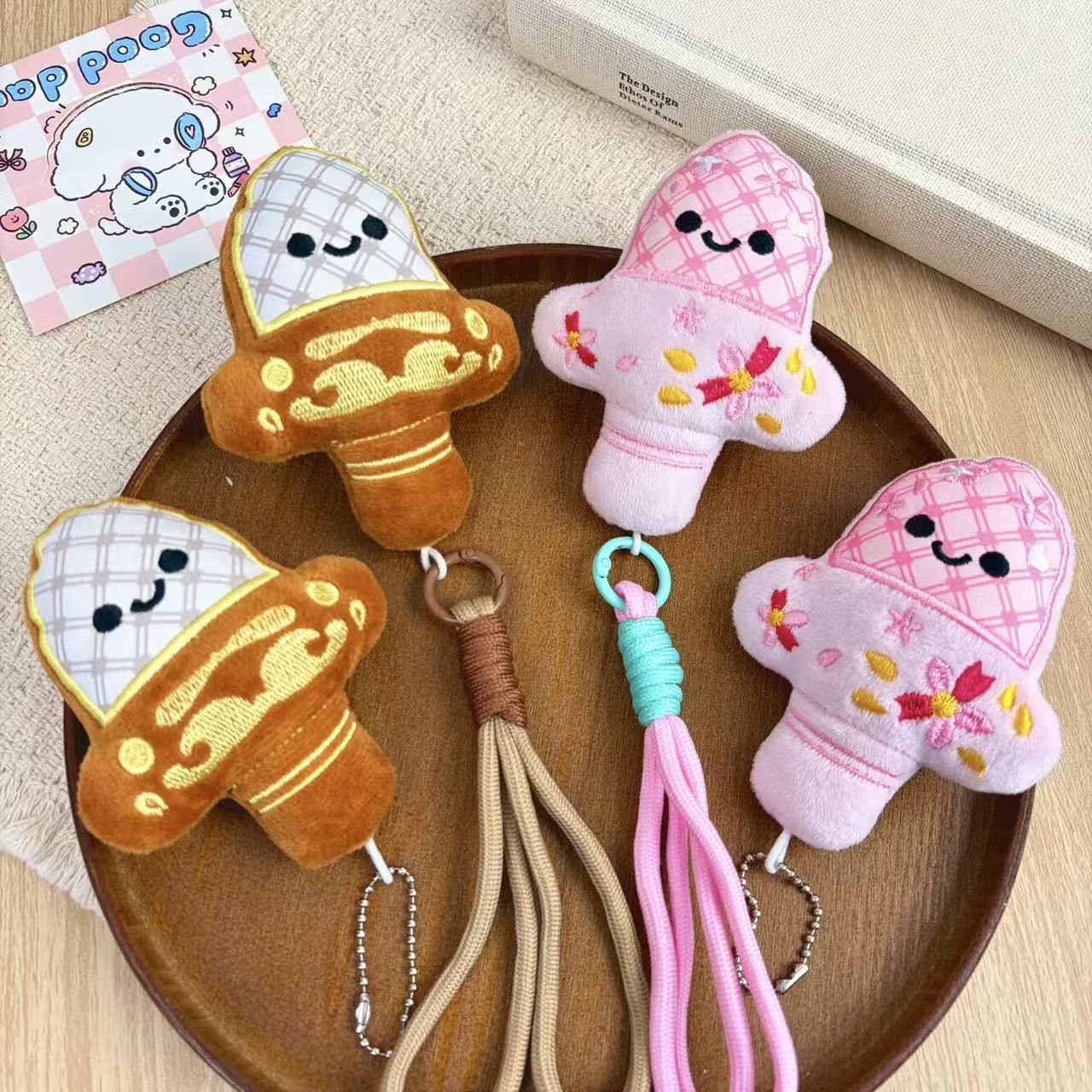 Cute mushroom plush keychains with braided wrist lanyards in pink and brown plaid patterns on a wooden tray.
