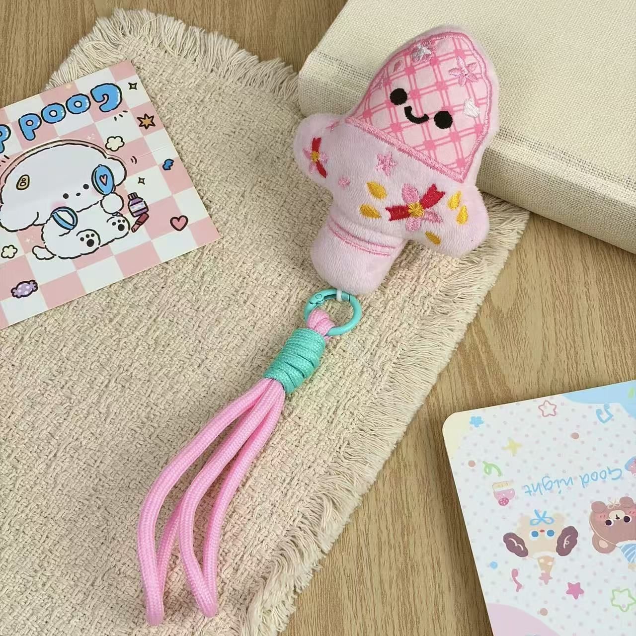 Pink plush mushroom keychain with an embroidered smiling face, plaid cap, floral details and a matching pink braided wrist strap on a cream cloth.