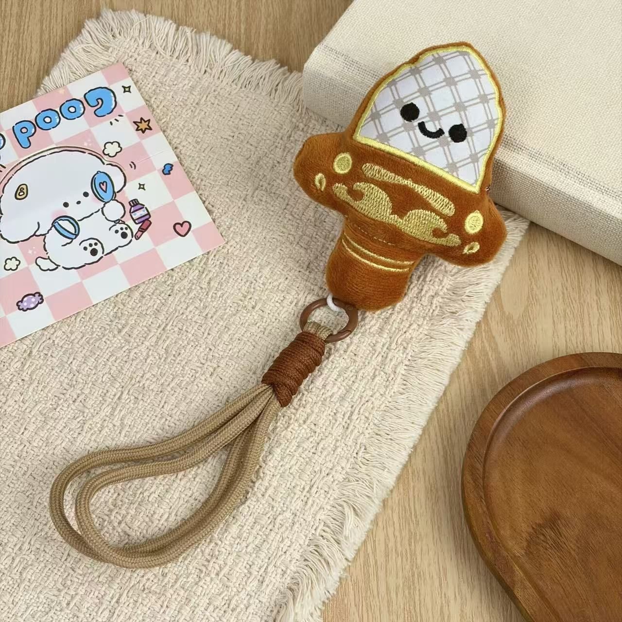 Brown plush mushroom keychain with embroidered smiling face and matching braided wrist strap on a cream cloth.